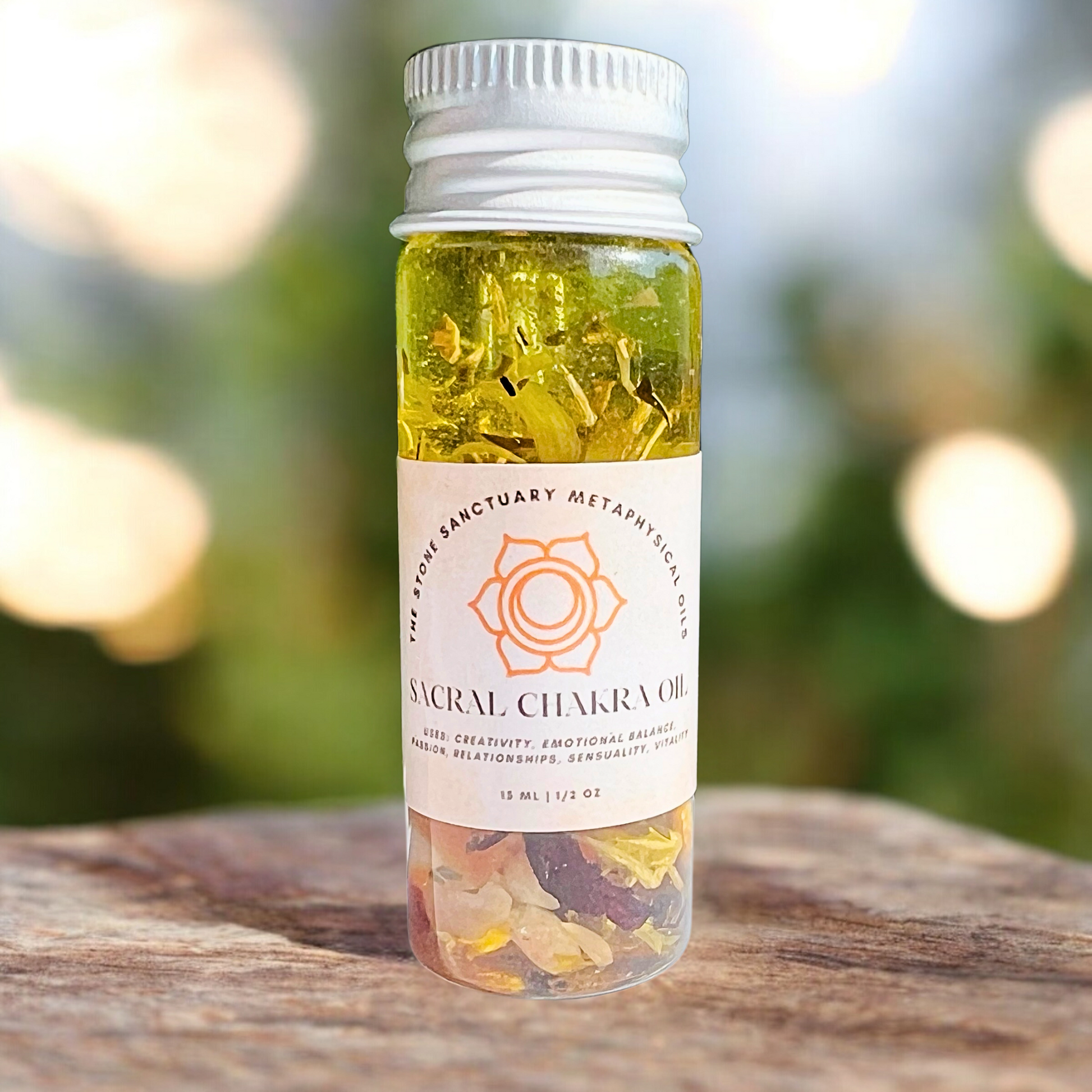 A small glass bottle of Sacral Chakra Oil with yellow liquid and tiny crystals bears a label featuring a geometric symbol and listing benefits like enhanced creativity, emotional balance, and improved relationships. The bottle is nestled amid green succulent plants.