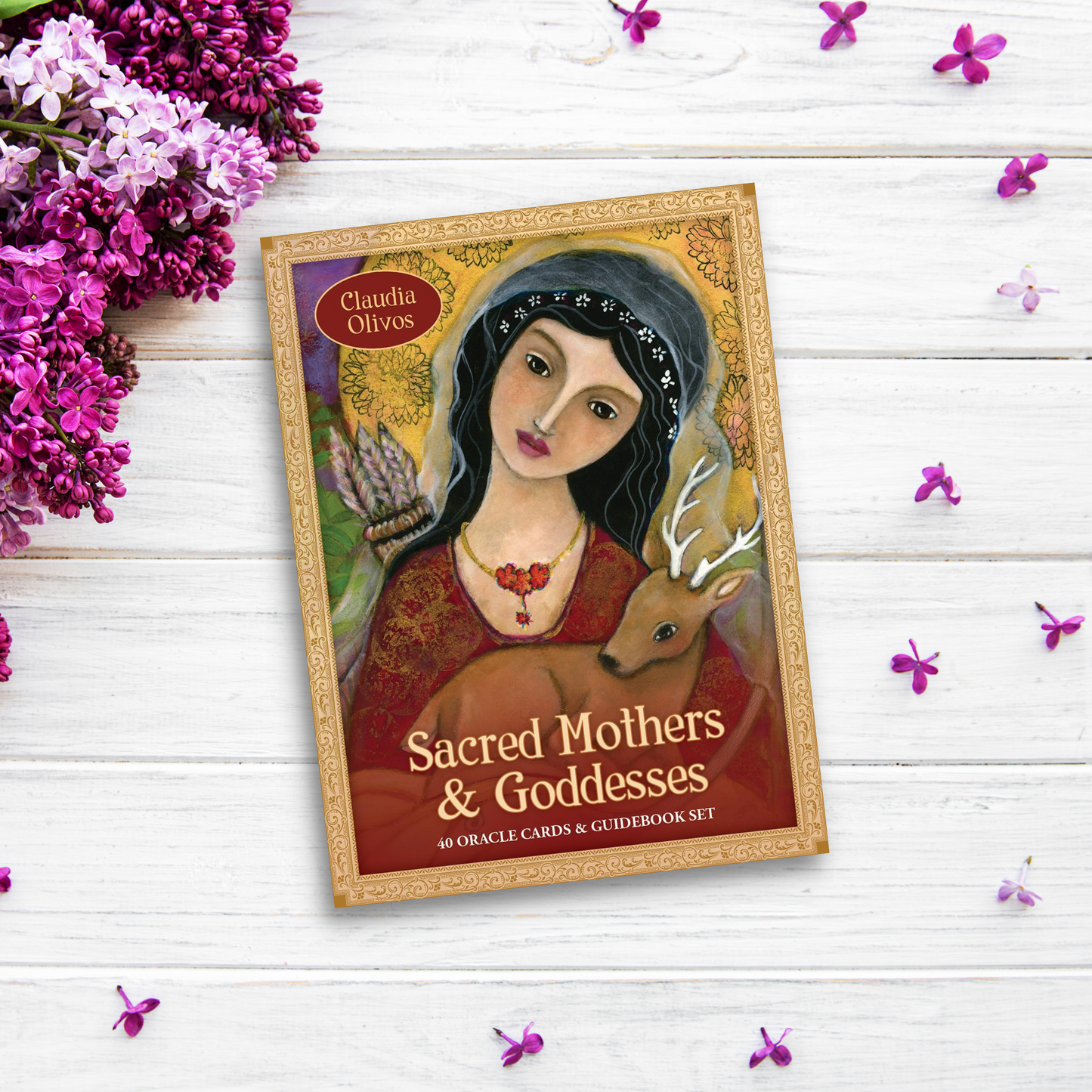 The image shows a goddess deck titled "Sacred Mothers and Goddesses: 40 Oracle Cards & Guidebook Set" by Claudia Olivos. The box features an illustrated woman with a deer, wearing a floral headpiece and red necklace. It is placed on a white wooden surface surrounded by purple and white flowers, exuding sacred feminine energy.