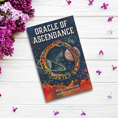 A copy of "Oracle of Ascendance" by Phillip J. Adamczyk and Sheri Jo Posselt lies on a white wooden surface surrounded by purple flowers. The cover features cosmic artwork with a snake encircling planets against a starry background, hinting at the metaphysical systems explored within.