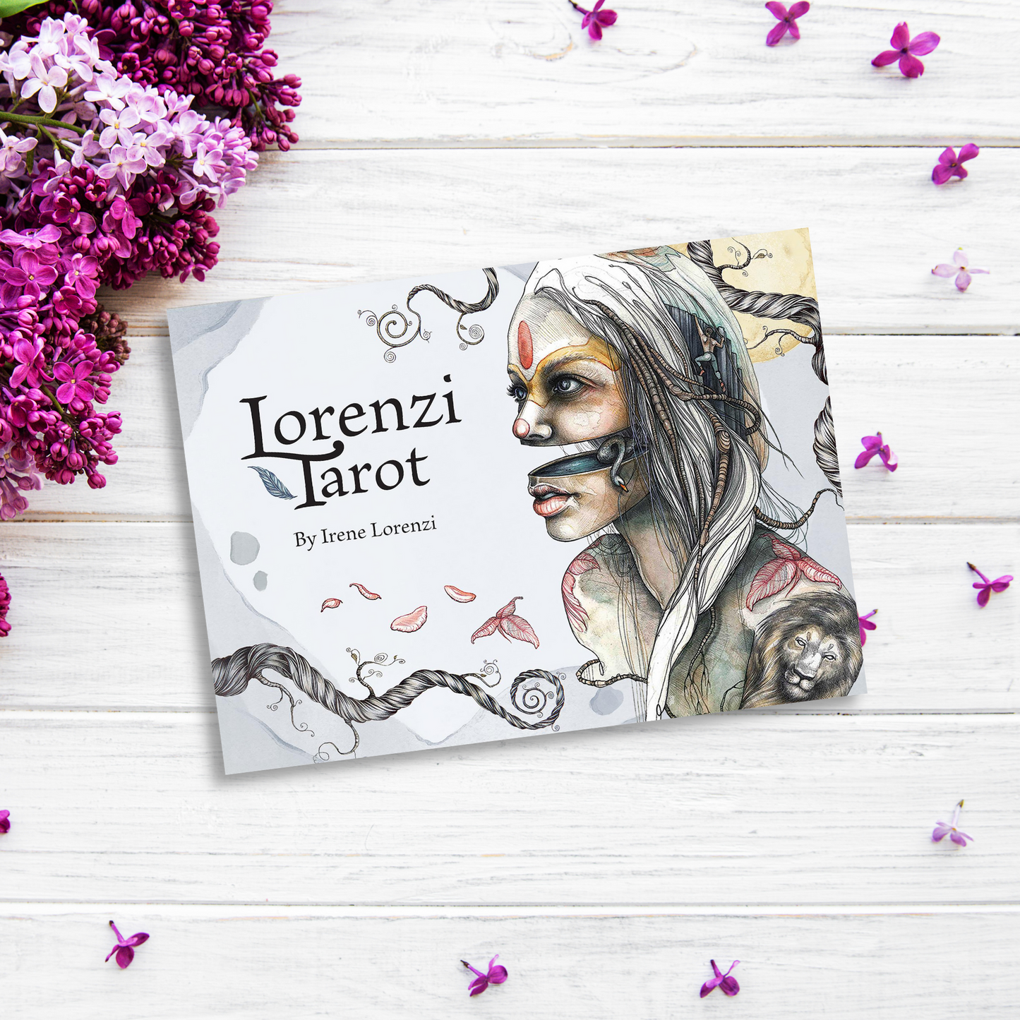 An image of Lorenzi Tarot positioned on a white wooden surface. The tarot deck card features an intricate illustration of a woman with tribal face paint and accessories. Surrounding the card are clusters of pink-purple flowers and scattered petals, capturing the essence of dualism.