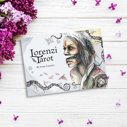 An image of Lorenzi Tarot positioned on a white wooden surface. The tarot deck card features an intricate illustration of a woman with tribal face paint and accessories. Surrounding the card are clusters of pink-purple flowers and scattered petals, capturing the essence of dualism.