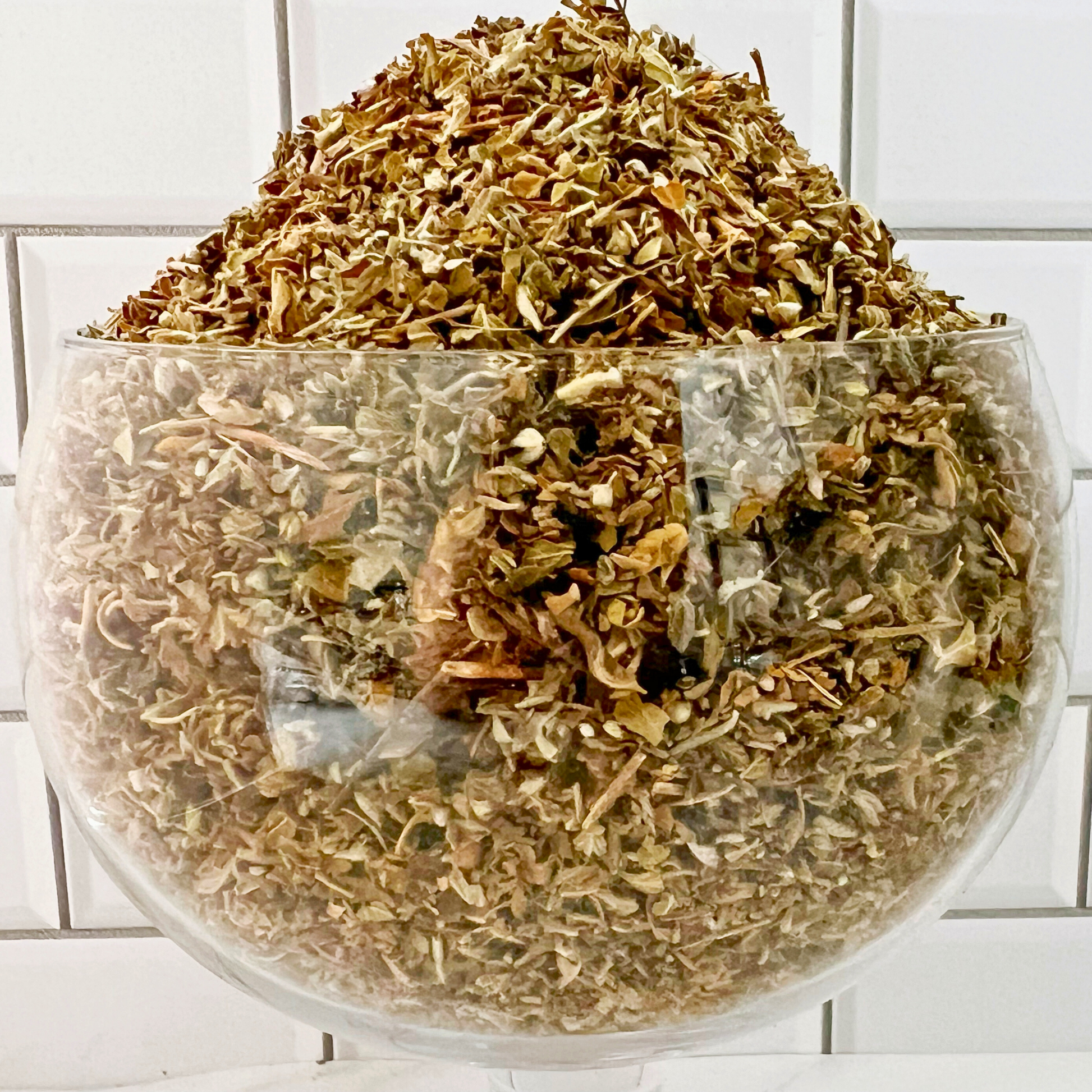 A bag labeled "Damiana Leaf" is placed in front of a glass bowl filled with Damiana Leaf. To the right, a white mortar and pestle are visible. This setup is against a white tiled background, suggesting uses such as enhancing dreams, work, love, energy, and other metaphysical practices.