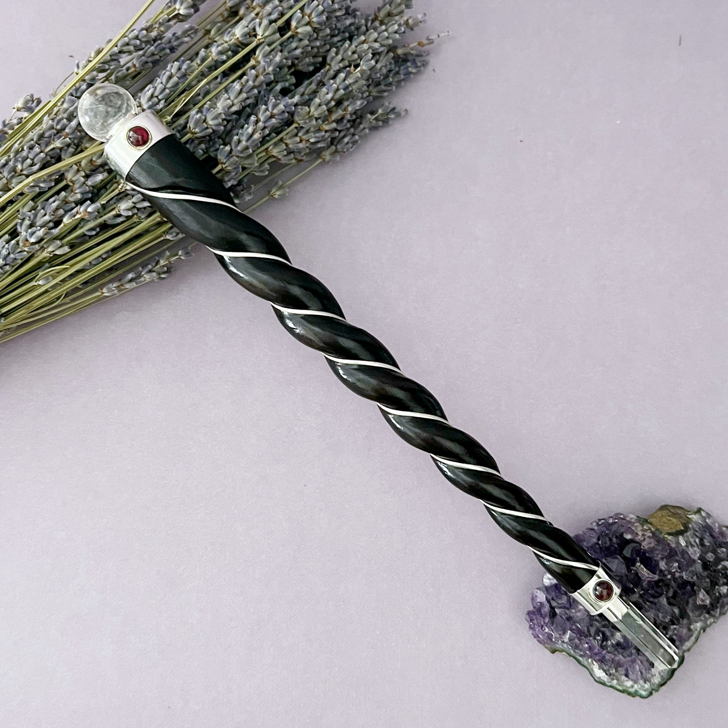 A Twisted Rosewood & Quartz Healing Wand, featuring a black, spiral-wrapped design with a clear crystal at the top and red gems on the handle, rests on a light purple surface. It is positioned diagonally among lavender sprigs and an amethyst cluster, making it ideal for spiritual practices.