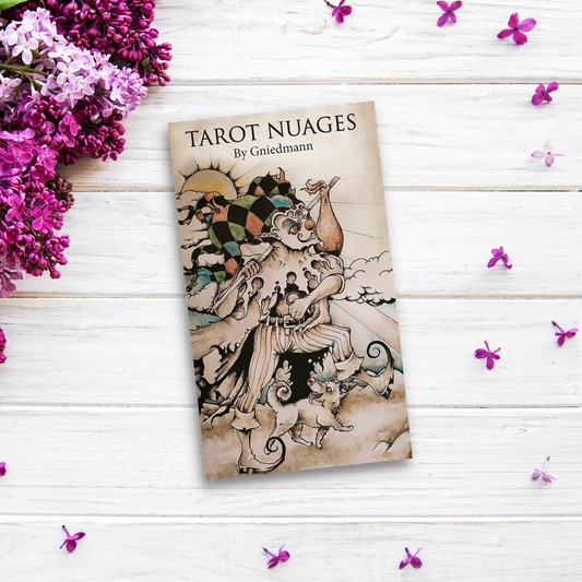 A book titled "Tarot Nuages" by Gniedmann lies on a white wooden surface, surrounded by small purple flowers and a bunch of lilacs. The cover features an artistic, mystical illustration inspired by Marseilles-style tarot, with intricate depictions of the Major Arcana.