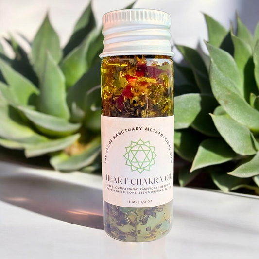 A small glass bottle labeled "Heart Chakra Oil" contains liquid and assorted herbs, designed to promote emotional healing. Positioned against a backdrop of green succulent plants, the bottle features a white cap and a geometric heart chakra symbol on the label, symbolizing the essence of unconditional love.