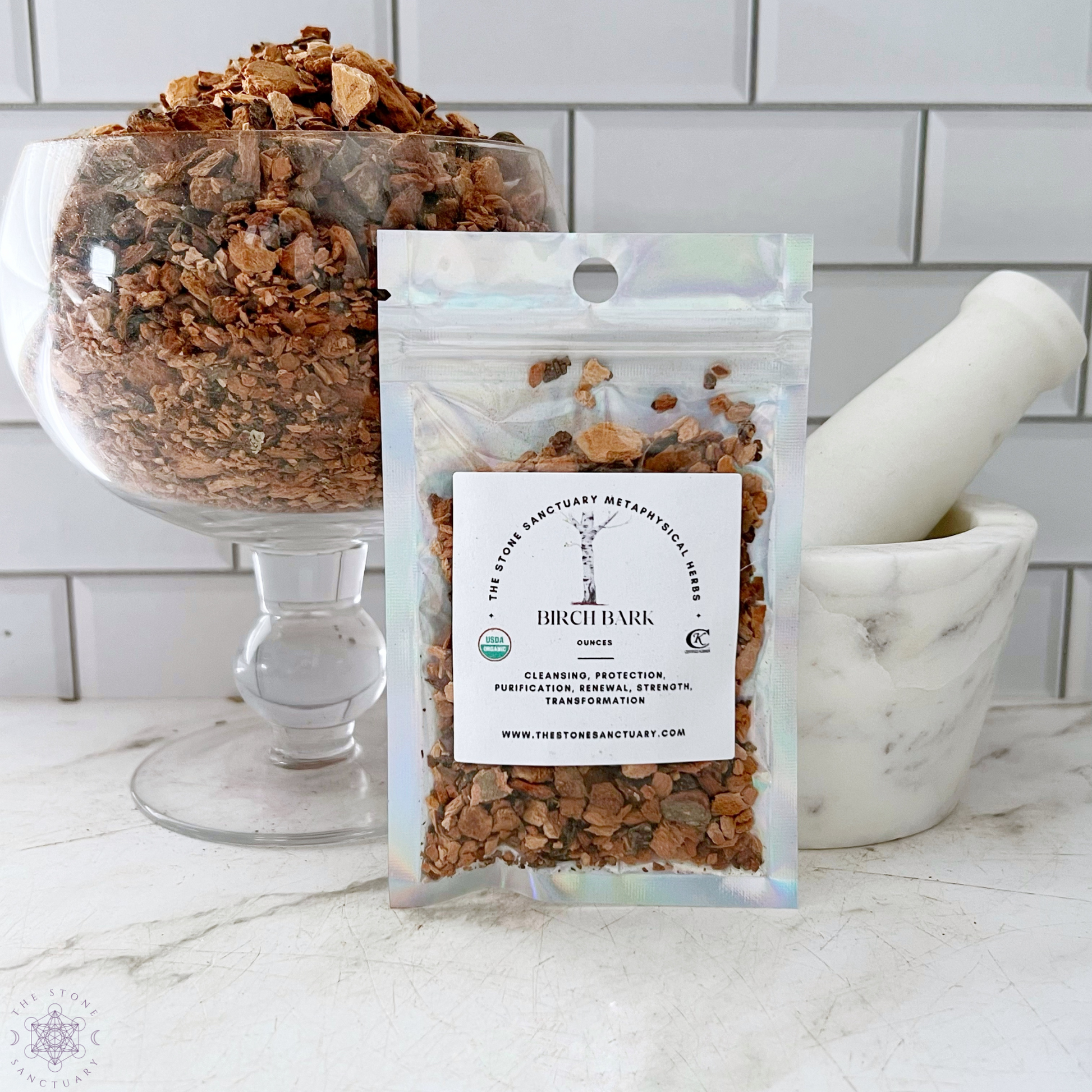 A small, holographic bag of Birch Bark, labeled "Birch Bark - Betula pubescens," is displayed in front of a larger glass bowl filled with birch bark pieces. A white marble mortar and pestle sit beside the bag. The background features a white tiled wall, highlighting its potential metaphysical uses.