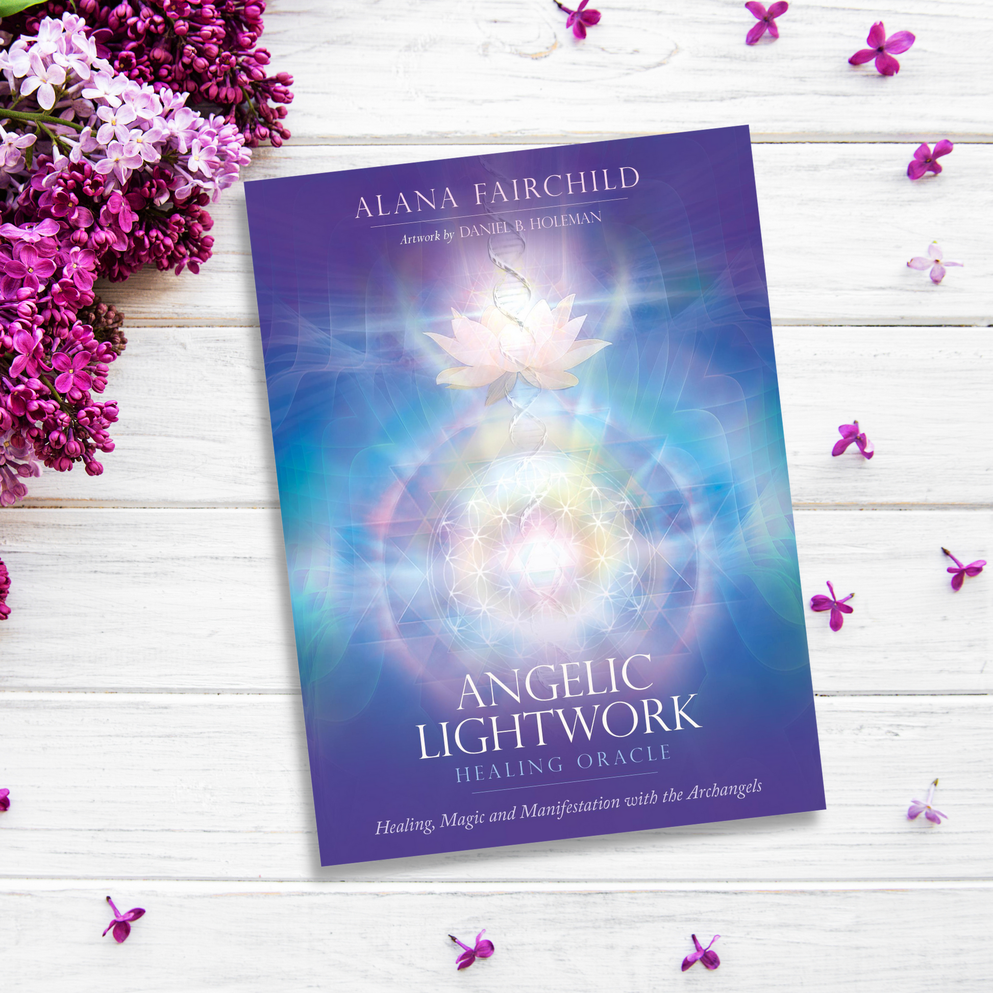 A book titled "Angelic Lightwork Healing Oracle" by Alana Fairchild, featuring artwork by Daniel B. Holeman, rests on a white wooden surface. The cover displays a luminous, mystical design with a lotus flower. Lilac flowers and petals surround the book, inviting spiritual awakening and healing practices.