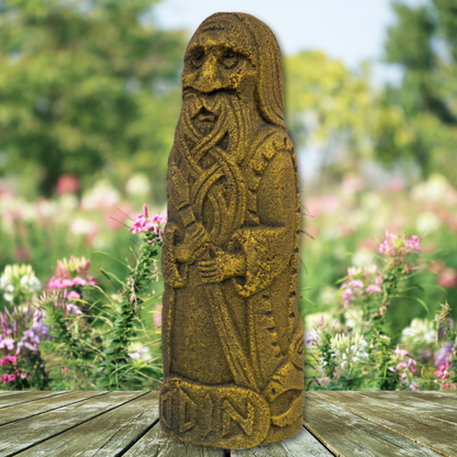 A weathered Odin Volcanic Statue stands on a wooden platform. The elderly bearded figure holding a staff is set against a vibrant, sunlit garden with blooming flowers and lush greenery, evoking the essence of Norse mythology.