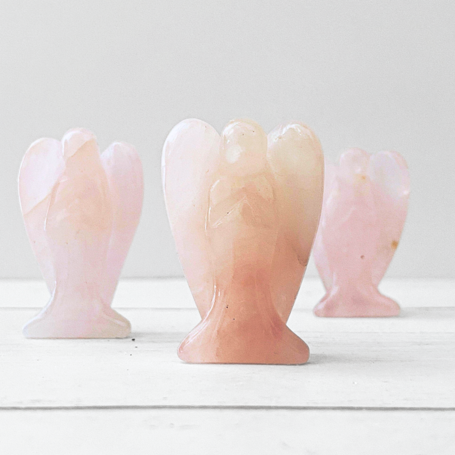 A polished pink rose quartz stone of unconditional love carved into the shape of an angel. The figure has wings extending slightly behind and a simple, abstract form without detailed facial features. This 1.5" Mini Rose Quartz Crystal Angel boasts a smooth, shiny surface with subtle variations in pink tones.