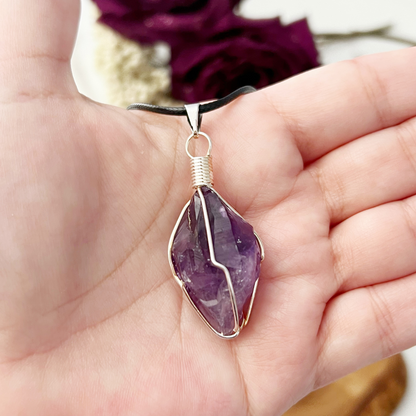 The 1" Wire Wrapped Raw Amethyst Crystal Point Pendant showcases a polished amethyst stone beautifully encased in intricately wrapped silver wire. Suspended on a black cord, the pendant features a purple amethyst with varying shades and visible facets, giving it an authentic, natural appearance. Known for its healing properties, this pendant offers both elegance and charm.