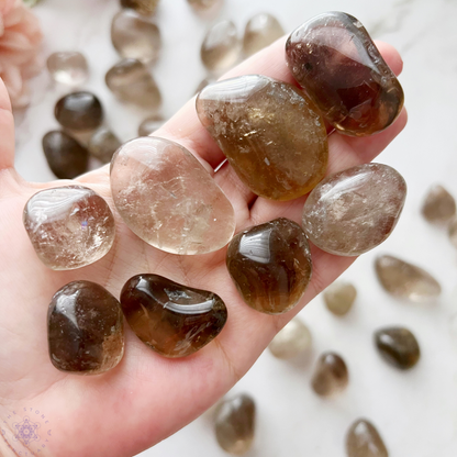 A wooden bowl filled with smooth, polished Smoky Quartz Tumbled Stones sits against a plain white background. The stones, known for their grounding stone qualities and healing properties, vary in shades of translucent gray and brown, with natural inclusions and cracks visible within them.