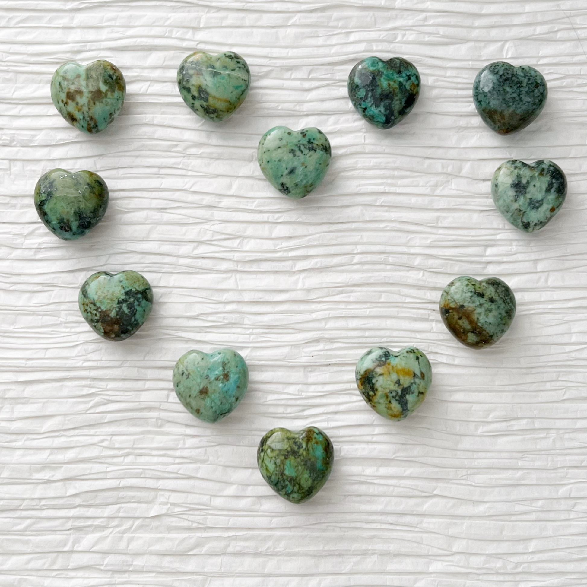 1" Mini African Turquoise Hearts are displayed, with some resting on a white, round, translucent surface and others on a gold dish. Green foliage is partially visible in the background. The heart-shaped gemstones feature a mix of green and darker shades, creating a natural pattern.