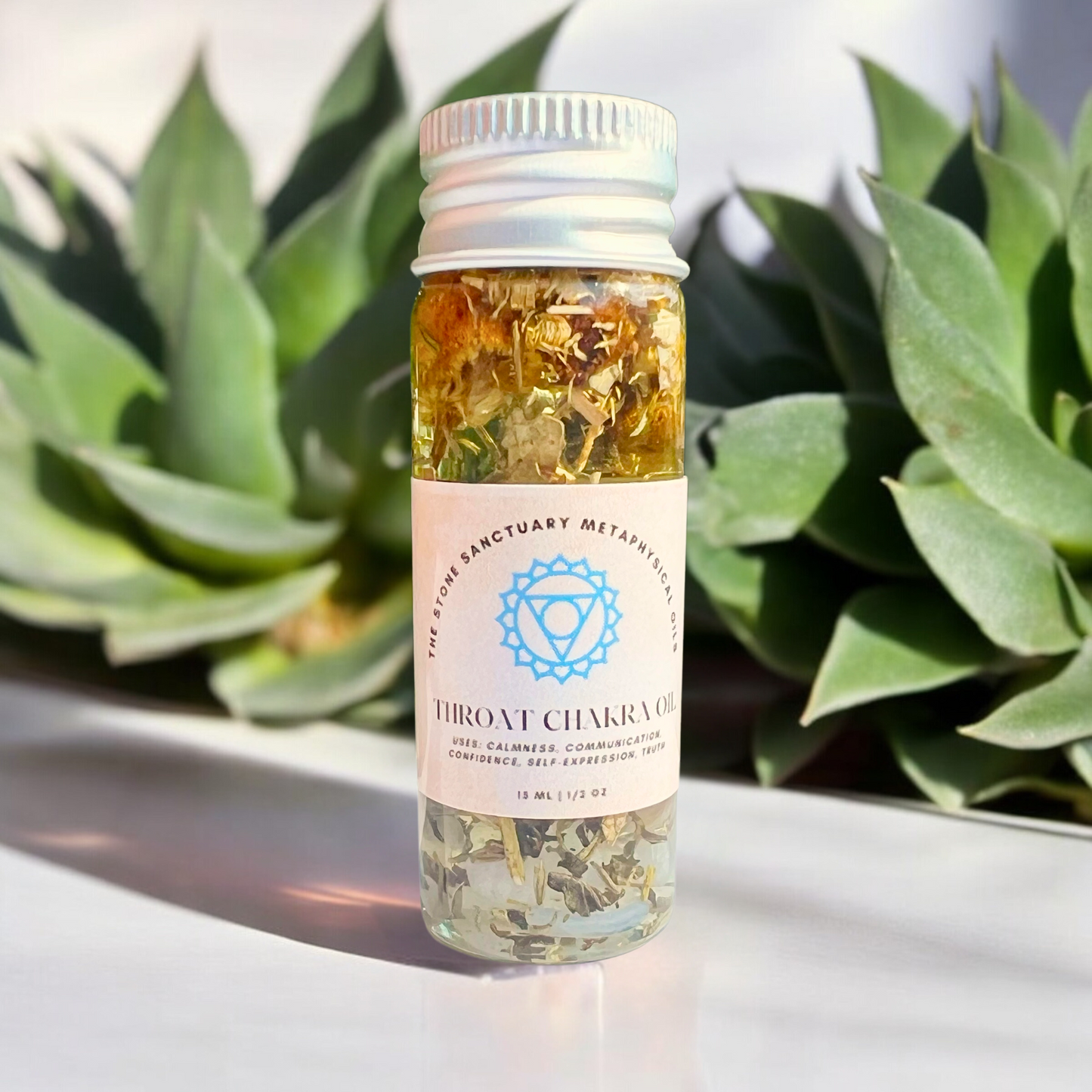 A small bottle of "Throat Chakra Oil," filled with a herbal mixture, sits on a white surface with a white cap. The blue design symbolizes the throat chakra and promotes self-expression. Blurred green succulent plants in the background enhance the setting for authentic communication.