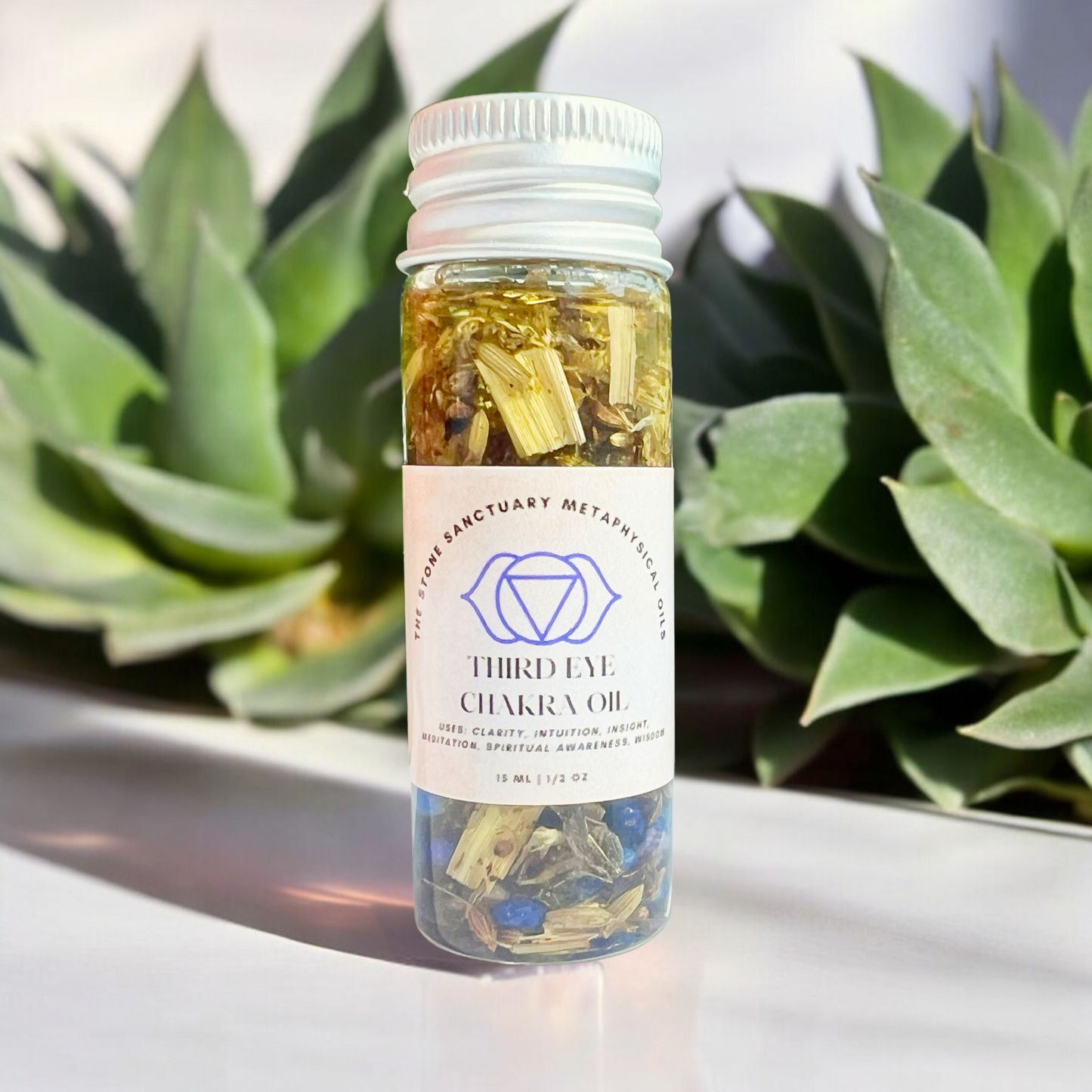 A small, clear bottle labeled "Third Eye Chakra Oil" with yellow and blue crystals inside sits gracefully in front of green succulent plants, the focus entirely on the bottle. The soft, blurred background accentuates the essence of spiritual awareness encapsulated within.