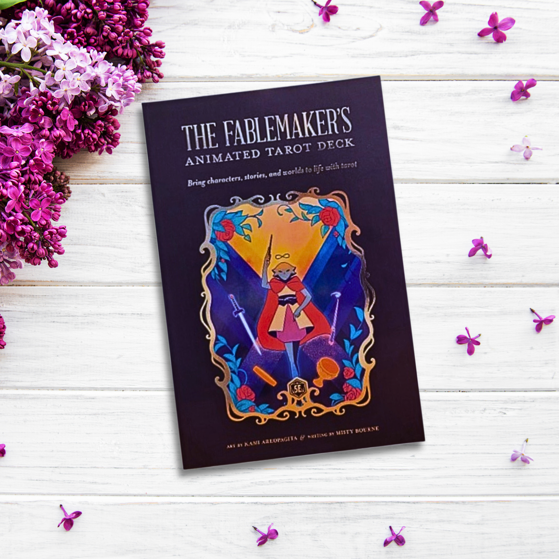 A book titled "The Fablemaker's Animated Tarot Deck" with colorful graphics is placed on a white wooden surface. Surrounding The Fablemaker's Animated Tarot Deck are purple flowers and small pink petals, adding a vibrant and artistic touch to the composition, perfect for interactive tarot readings or enhancing your TTRPGs.