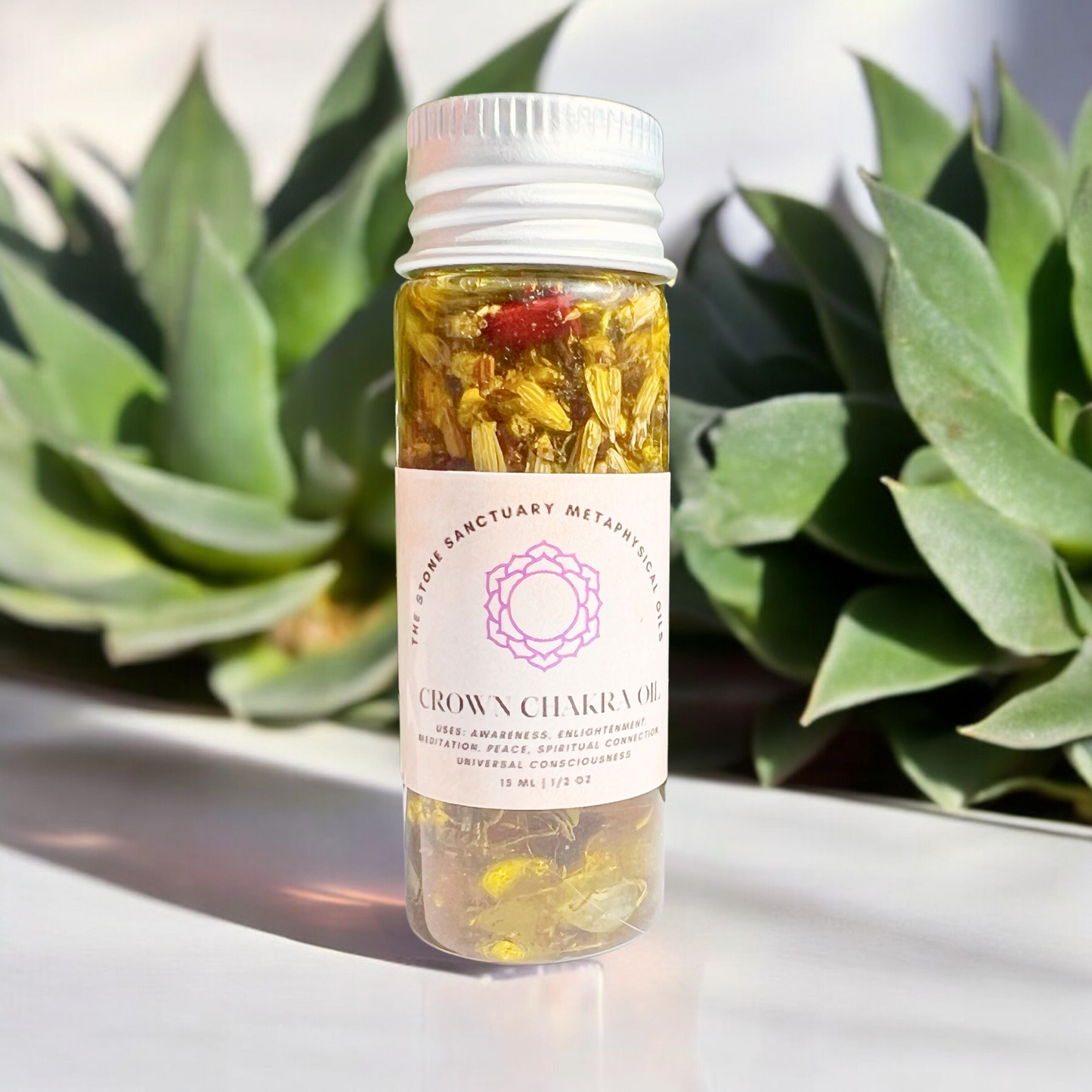 A small, transparent bottle with a white cap houses yellow flower petals and a red bud, labeled "Crown Chakra Oil" and adorned with a purple chakra symbol. Green leaves in the blurred background enhance the serene aura of this potent formula for divine connection, all beautifully set on a pristine white surface.