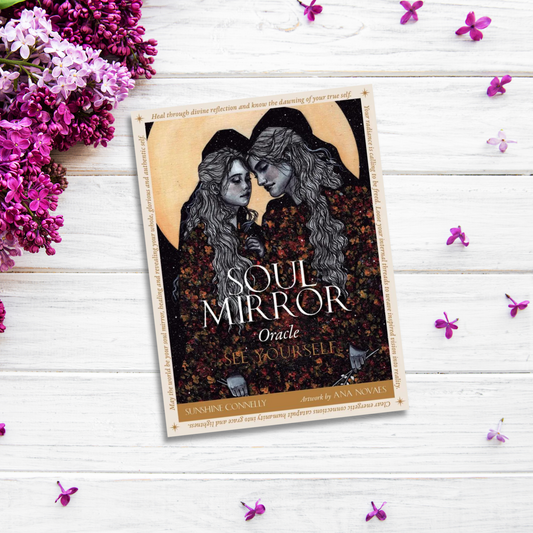 A product titled "Soul Mirror Oracle" by Sunshine Connolly, illustrated by Ana Novais, lies on a white wooden surface surrounded by clusters of purple flowers. This guidebook's cover features an illustration of two women with flowing hair, wearing dark clothing.