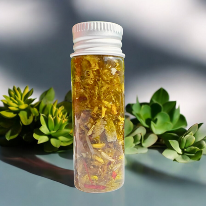 A small, transparent bottle with a white cap houses yellow flower petals and a red bud, labeled "Crown Chakra Oil" and adorned with a purple chakra symbol. Green leaves in the blurred background enhance the serene aura of this potent formula for divine connection, all beautifully set on a pristine white surface.