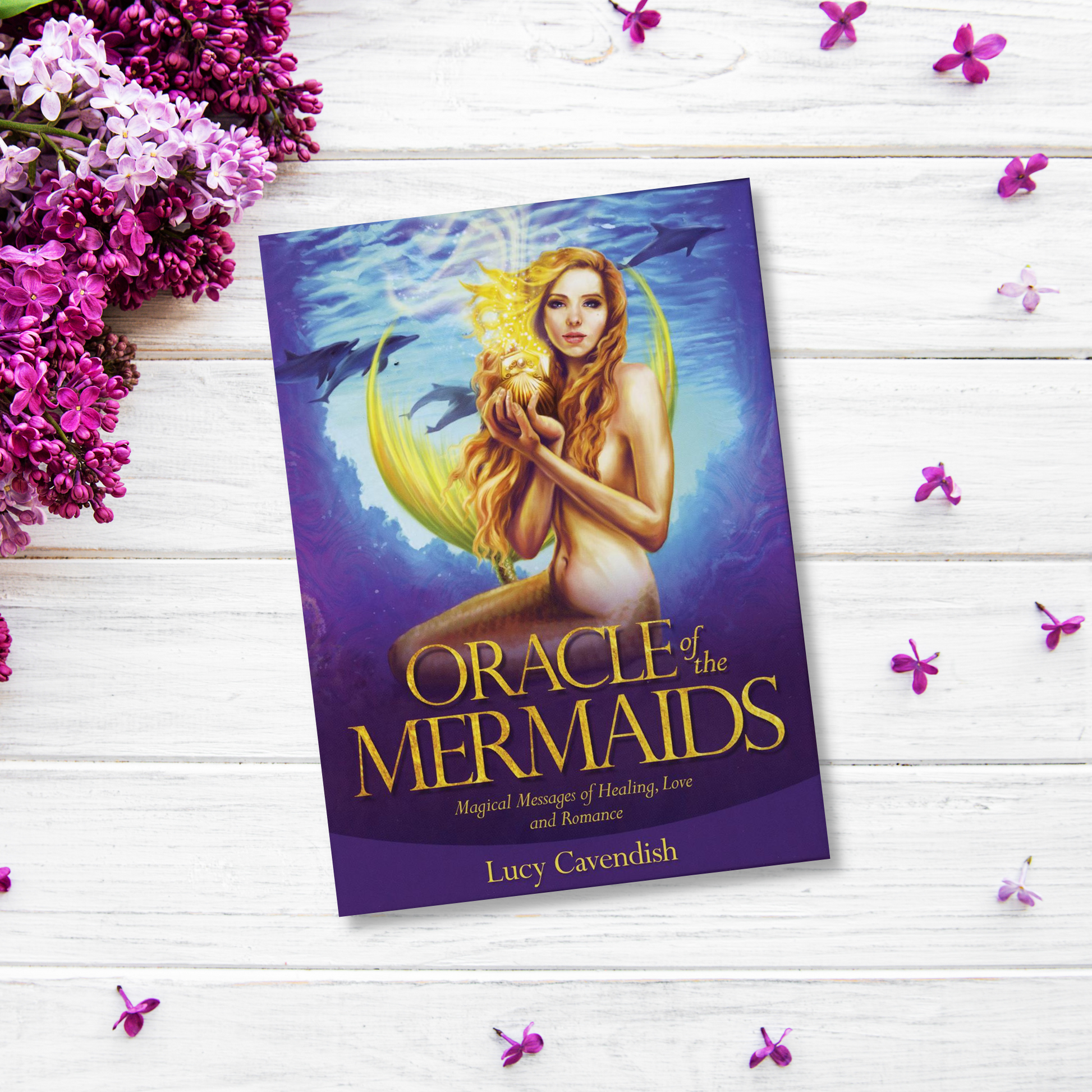 The product "Oracle of the Mermaids" by Lucy Cavendish is placed on a white wooden surface, surrounded by purple and white flowers. The cover features a mermaid holding a glowing orb, embodying Mermaid Magic and the Feminine Divine, with marine creatures in the background.