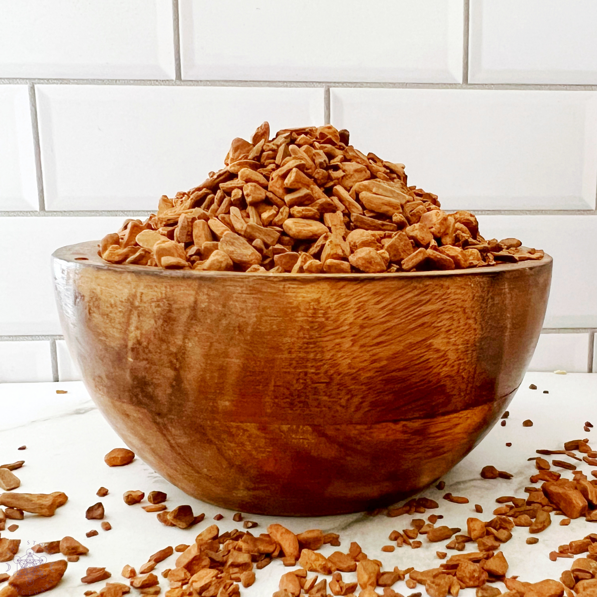 A small silver bag labeled "Cinnamon Chips" with information on the front stands next to a white mortar and pestle. Behind them is a large glass bowl filled with cinnamon bark pieces, perfect for enhancing your culinary endeavors. The background features a white tiled wall.