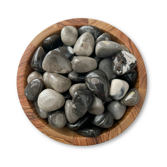 A wooden bowl filled with smooth, polished Flint Tumbled Stones of varying shades of black and gray graces a white background, offering both visual appeal and intriguing metaphysical properties.