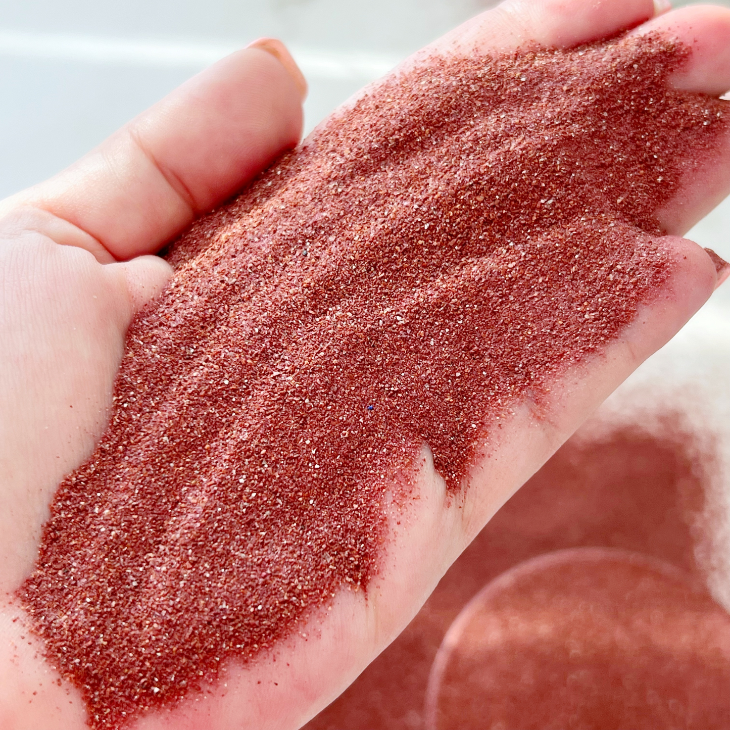 A small black bowl filled with Red Jasper Gemstone Sand, accompanied by two jars of Red Jasper Gemstone Sand on a white tiled surface. The open jar displays a lid labeled "Red Jasper" and a description of its properties, radiating peaceful energy.