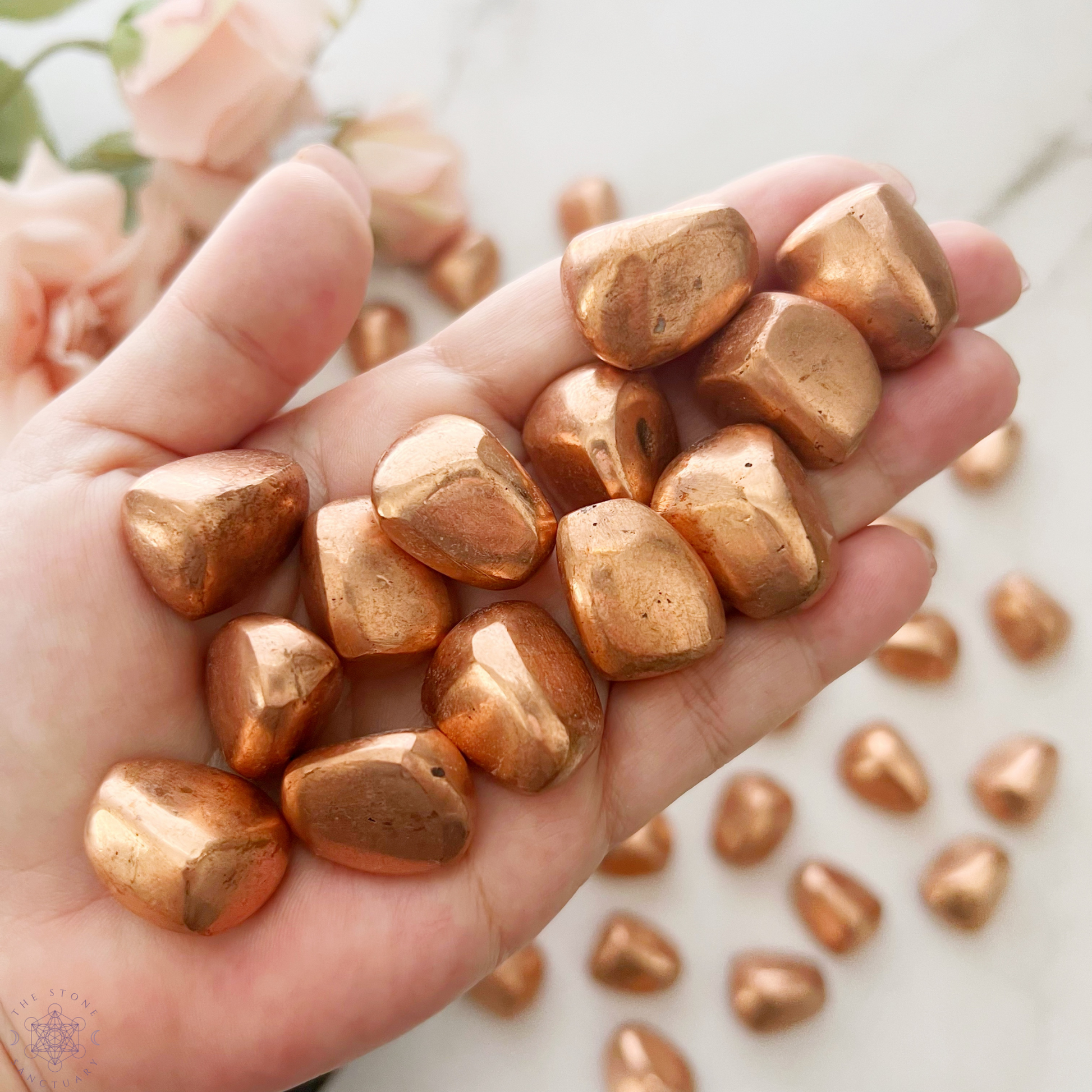 A wooden bowl filled with shiny, polished Copper Nugget Tumbled Stones is centered on a white background. These copper tumbled stones have a smooth, glossy finish and varying shapes, reflecting light to accentuate their metallic luster, perfect for chakra healing and energy transfer.