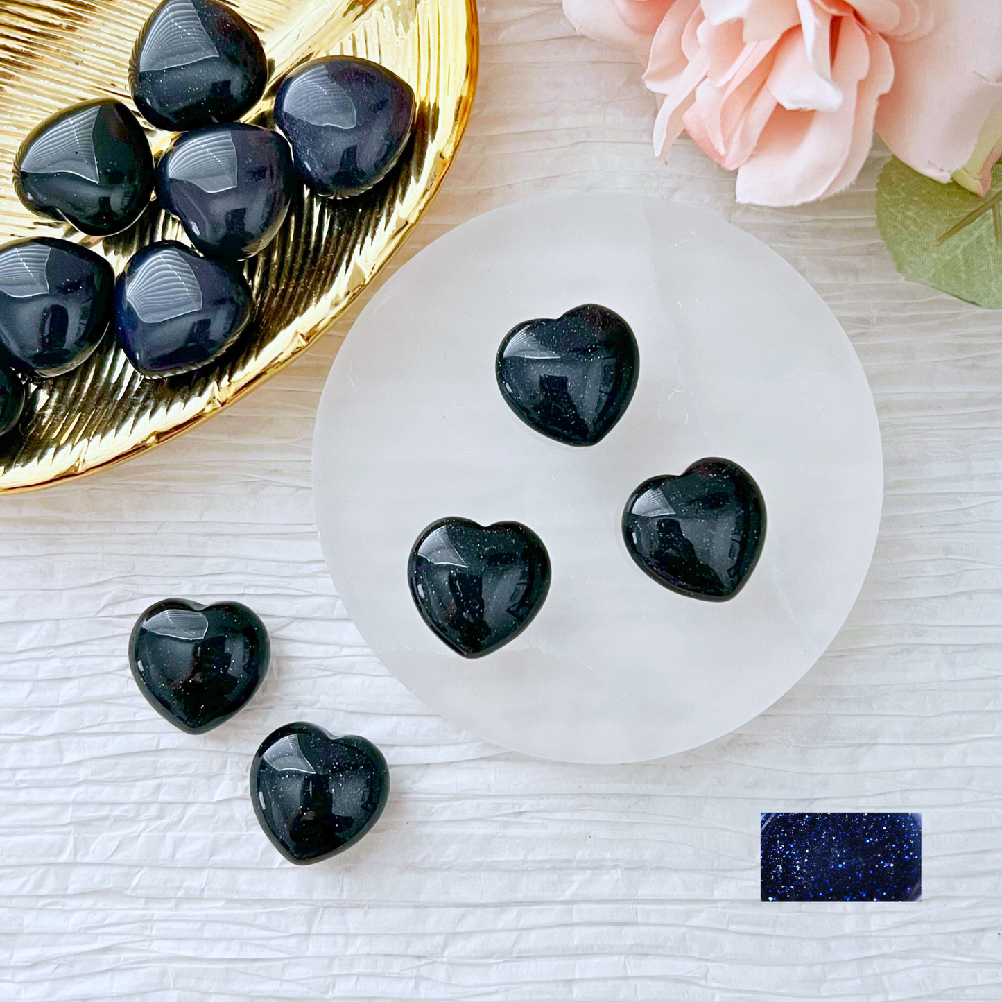 A marble surface displays small black heart-shaped stones. Four stones are arranged on a round white coaster, while others fill a gold heart-shaped dish. A dark blue rectangular 1" Mini Blue Goldstone Heart, known for enhancing spiritual perception and being a protective stone, and a light pink rose are also in the frame.