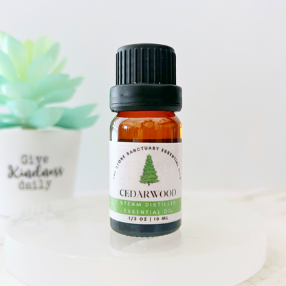 A small amber bottle of Cedarwood Essential Oil sits on a white surface. The bottle, featuring a black cap and a green and white label with a tree graphic, contains steam distilled essential oil. In the background, there is a potted succulent with a partially blurred quote: "Give Kindness daily.