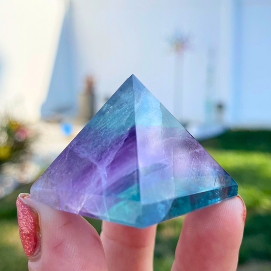 A hand with sparkly pink nail polish holds a transparent 1" Mini Rainbow Fluorite Pyramid with shades of blue, purple, and green. The background is an outdoor setting with soft-focus greenery and a light-colored building, perfect for mental and spiritual growth or to boost creativity.