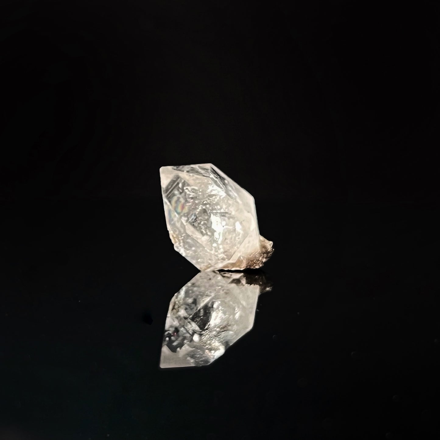 A single, .68" Raw Herkimer Diamond is shown against a black background. The unique, powerful crystal's reflective surface creates a mirrored image on the dark surface beneath it, highlighting its sharp edges and intricate internal structure.