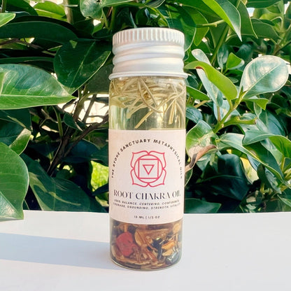 A petite clear bottle, labeled "Root Chakra Oil," is filled with dried herbs and delicate flowers. Featuring a white cap, the bottle is set against a backdrop of lush green foliage, crafting a natural and serene scene that inspires feelings of grounding and stability.