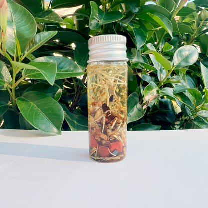 A petite clear bottle, labeled "Root Chakra Oil," is filled with dried herbs and delicate flowers. Featuring a white cap, the bottle is set against a backdrop of lush green foliage, crafting a natural and serene scene that inspires feelings of grounding and stability.