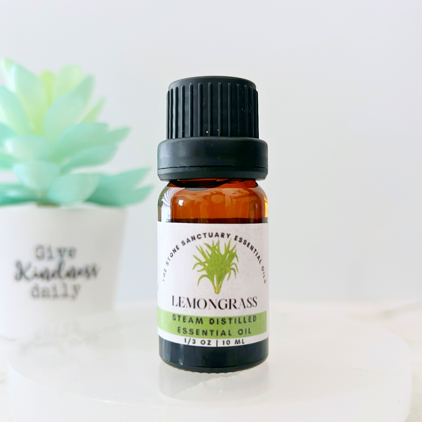 A small amber bottle with a black cap labeled "Lemongrass Essential Oil" rests on a white surface. In the background, there's a succulent plant in a white pot with the inscription "Give Kindness Daily." Experience the premium quality and soothing aroma of this exquisite oil.