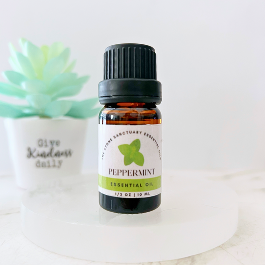 A small brown glass bottle of Peppermint Essential Oil with a black cap sits on a white surface. The bottle's label features a green peppermint leaf and the text "PEPPERMINT Essential Oil, 1/3 OZ 10ML." In the background, a potted succulent and a sign that reads "Give Kindness Daily" are visible. This steam-distilled, premium quality oil