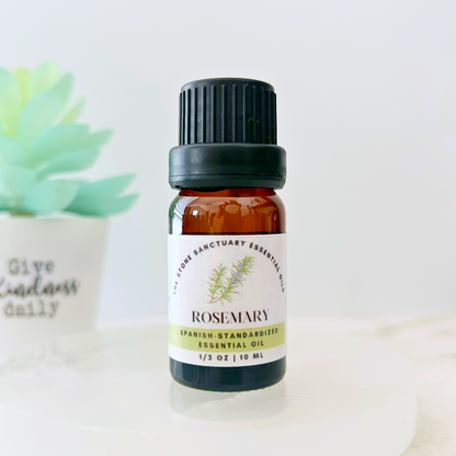 A small amber bottle of Rosemary Essential Oil with a black cap, labeled "Rosemary Spanish-Standardized Essential Oil 1/3 oz | 10 ml." A succulent plant and a white pot inscribed with "Give Kindness Daily" are blurred in the background. Steam distilled for purity.