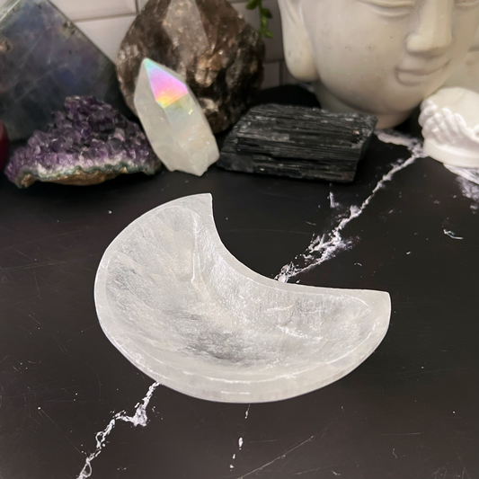 A 4" Selenite Crescent Moon Bowl is displayed on a black marble surface. Surrounding the bowl are various other crystals in different shapes and colors, along with a Buddha head statue.