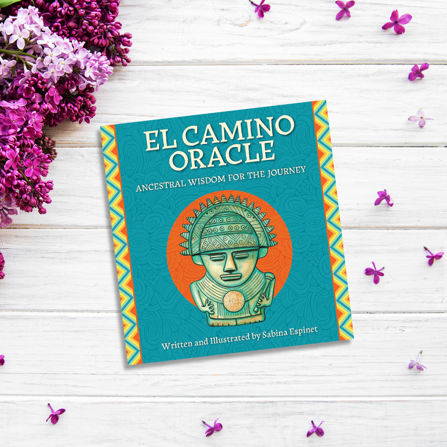 A book titled "El Camino Oracle" by Sabina Espinet is placed on a white wooden surface surrounded by purple flowers and green leaves. The cover, highlighting cultural heritage, features an illustration of a stylized figure with an intricate headdress on an orange background.