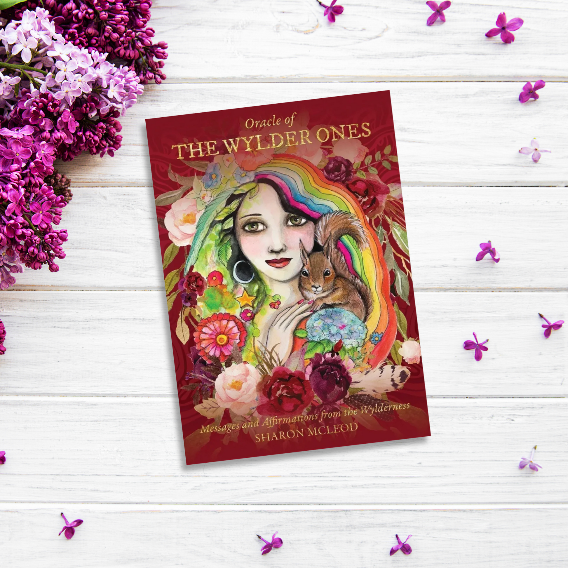 A book titled "Oracle of The Wylder Ones" by Sharon McLeod lies on a white wooden surface. The cover features a colorful illustration of a woman, flowers, and a squirrel. Purple and pink flowers surround the book, promoting self-awareness.