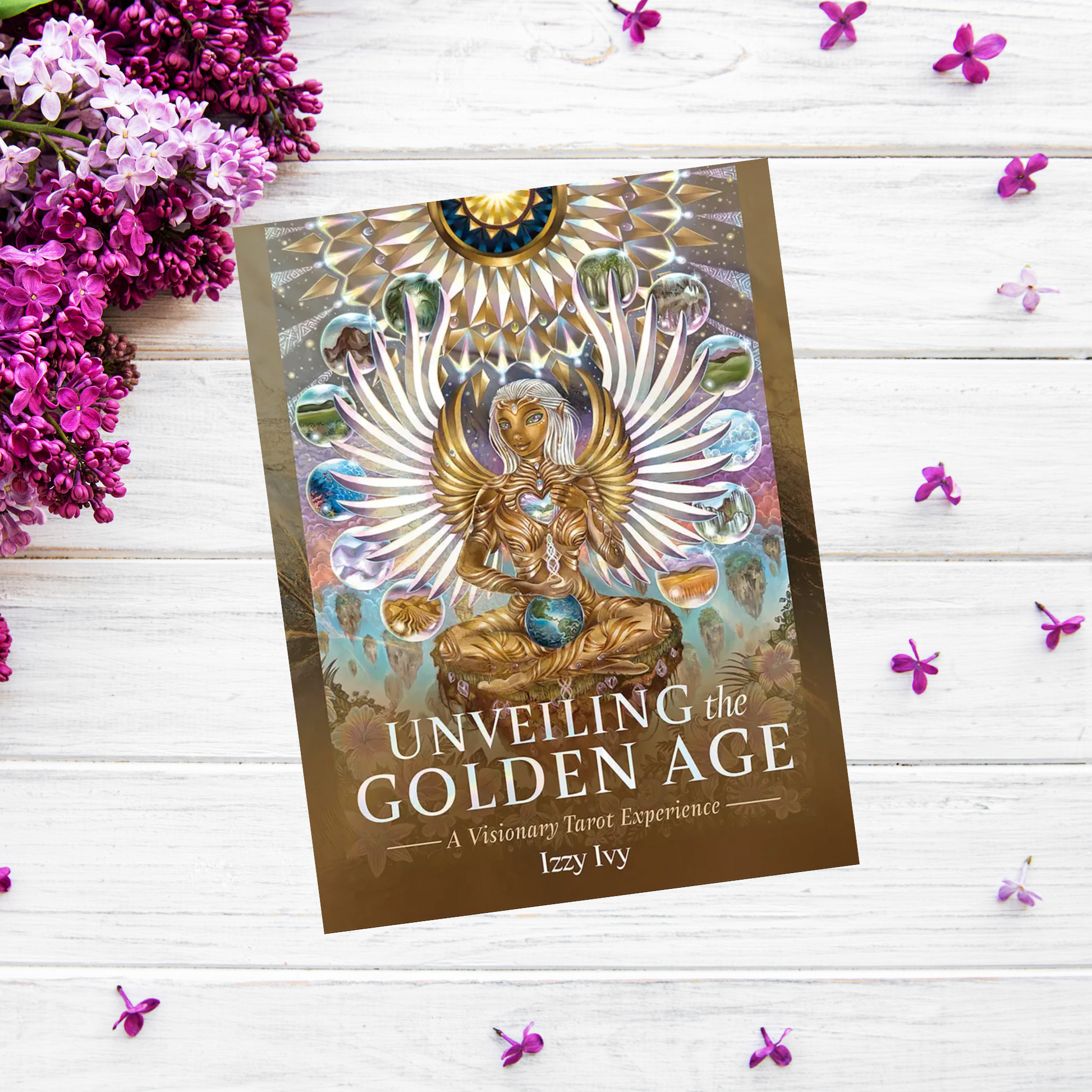 A product titled "Unveiling The Golden Age Tarot: A Visionary Tarot Experience" by Izzy Ivy lies on a white wooden surface adorned with scattered purple and white flowers. The book cover, reminiscent of a meditation deck, features an intricate illustration of a winged figure with ethereal elements and vibrant colors.