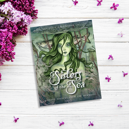 The image shows the book "Sisters Of The Sea" by Lucy Cavendish placed on a white wooden surface. The cover features an illustration of a green-haired mermaid. Purple flowers and scattered petals adorn the surface around the book, adding a touch of mermaid magic to the scene.