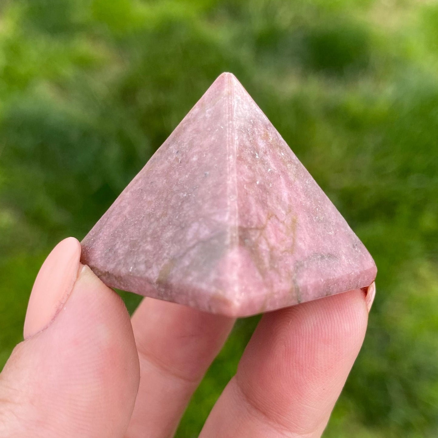 A hand holding a 1" Mini Rhodonite Pyramid against a blurred green grass background. The pyramid, a stunning addition to any crystal collection, has a smooth, glossy surface and distinct vertices, with subtle variations in pink hues and faint natural patterns.