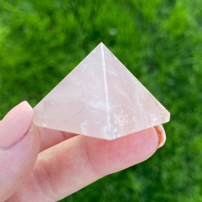 Three pink 1" Mini Rose Quartz Pyramids, known as the Stone of Unconditional Love, are displayed on a white wooden surface in front of a bundled sage smudge stick wrapped in string. The background features white subway tile, adding a clean and serene atmosphere to the composition.