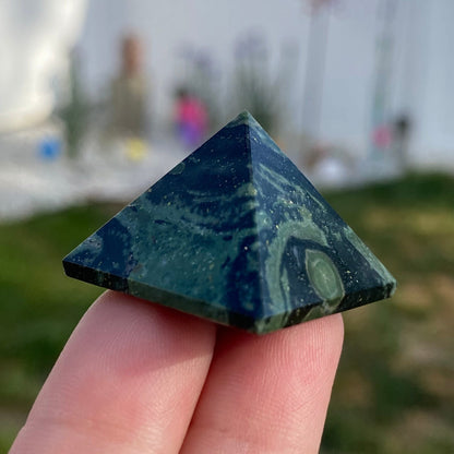 Three 1" Mini Kambaba Jasper Pyramids with calming green and black patterns are placed on a white surface. In the background, there is a bundle of sage tied with string, set against a white tiled wall. The scene is well-lit and has a clean, minimalist aesthetic.