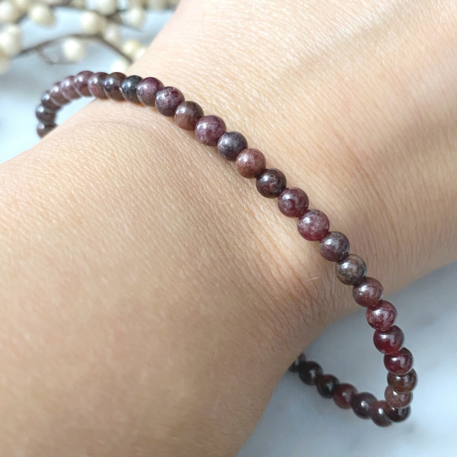 A stack of polished, 4mm Garnet Beaded Bracelets is displayed on a light surface. In the background, there is a blurred decorative element with white beads. The garnet beads are smooth and glossy, giving the bracelets an elegant appearance infused with Usui Reiki energy.