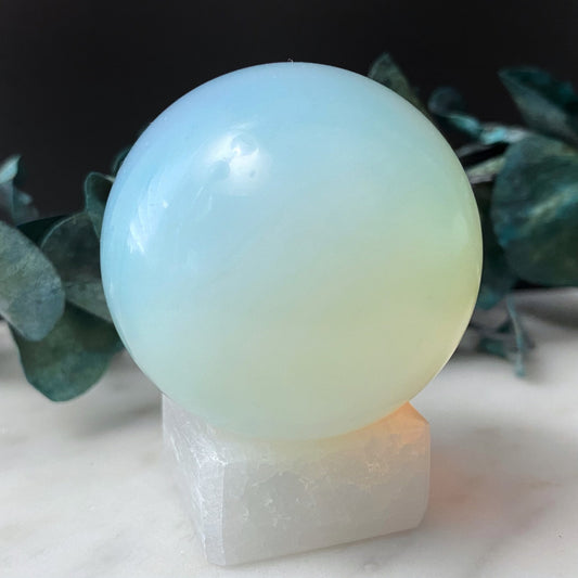 A smooth, polished 40mm Opalite Crystal Ball sits on a white, square base. The sphere has a luminous, milky appearance with soft gradients of blue and green. Perfect for use as a third eye chakra meditation tool, in the blurred background, green eucalyptus leaves are partially visible against a dark backdrop.