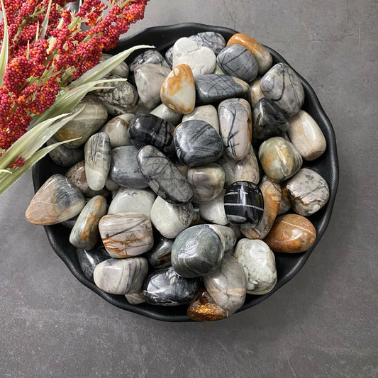 A black bowl filled with polished Picasso Jasper Tumbled Stones in shades of gray, white, black, and brown sits on a gray surface. Next to the bowl, a bunch of red and green plant stems adds a touch of creativity and texture to the setting.