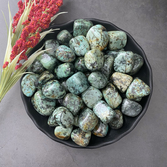 A black bowl filled with polished, light green and dark green speckled stones, known as African Turquoise Tumbled Stones, sits on a gray surface. To the left, a bundle of red berries and green leaves enhances the scene. These stones are often revered as the Stone of Evolution for their connection to Third Eye Wisdom.