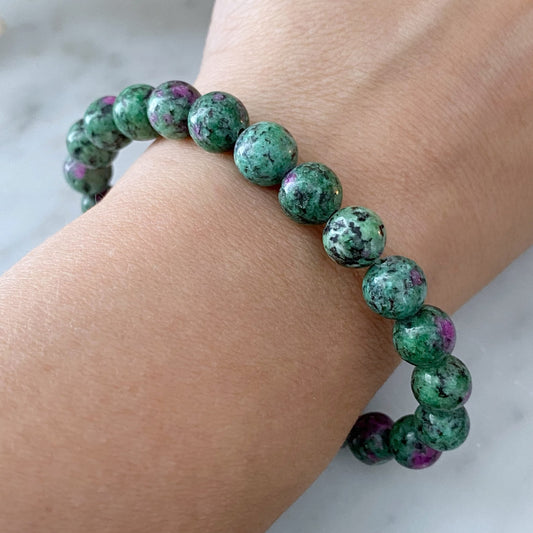 A close-up photo of a wrist adorned with an 8mm Ruby Zoisite Beaded Bracelet, showcasing green and purple speckled stones. The round, evenly sized beads create a simple yet elegant look against the light skin tone of the person's wrist. The background is a light, blurred surface, enhancing its subtle beauty.