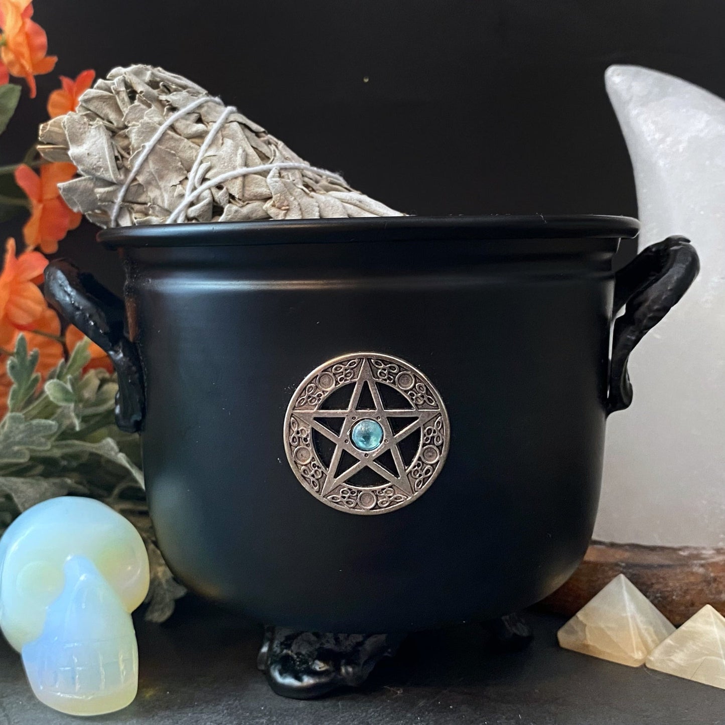 A black 4.5" Pentacle Metal Cauldron adorned with a pentagram emblem is filled with bundled sage. Surrounding the cauldron are an opalescent skull, a large white crystal, orange flowers, sage leaves, and small pyramid-shaped crystals, all set against a dark background.